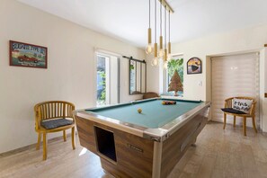 A pool table with a wooden finish and a glass top, surrounded by two wooden chairs with cushions.