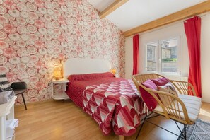 Un dormitorio con un papel tapiz estampado, una cama con colcha roja, una silla de madera y una mesita de noche con lámpara.