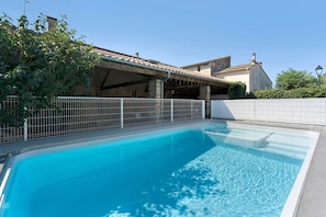 Una piscina con borde de baldosas, cerca de metal y un edificio con tejado de azulejos al fondo.