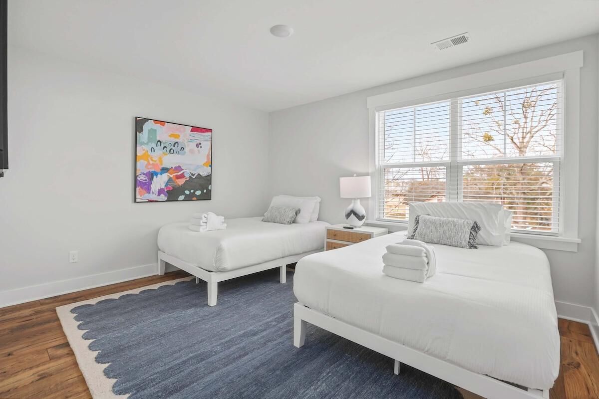 A bedroom with two white beds, a gray rug, a colorful abstract painting, and a window with blinds.