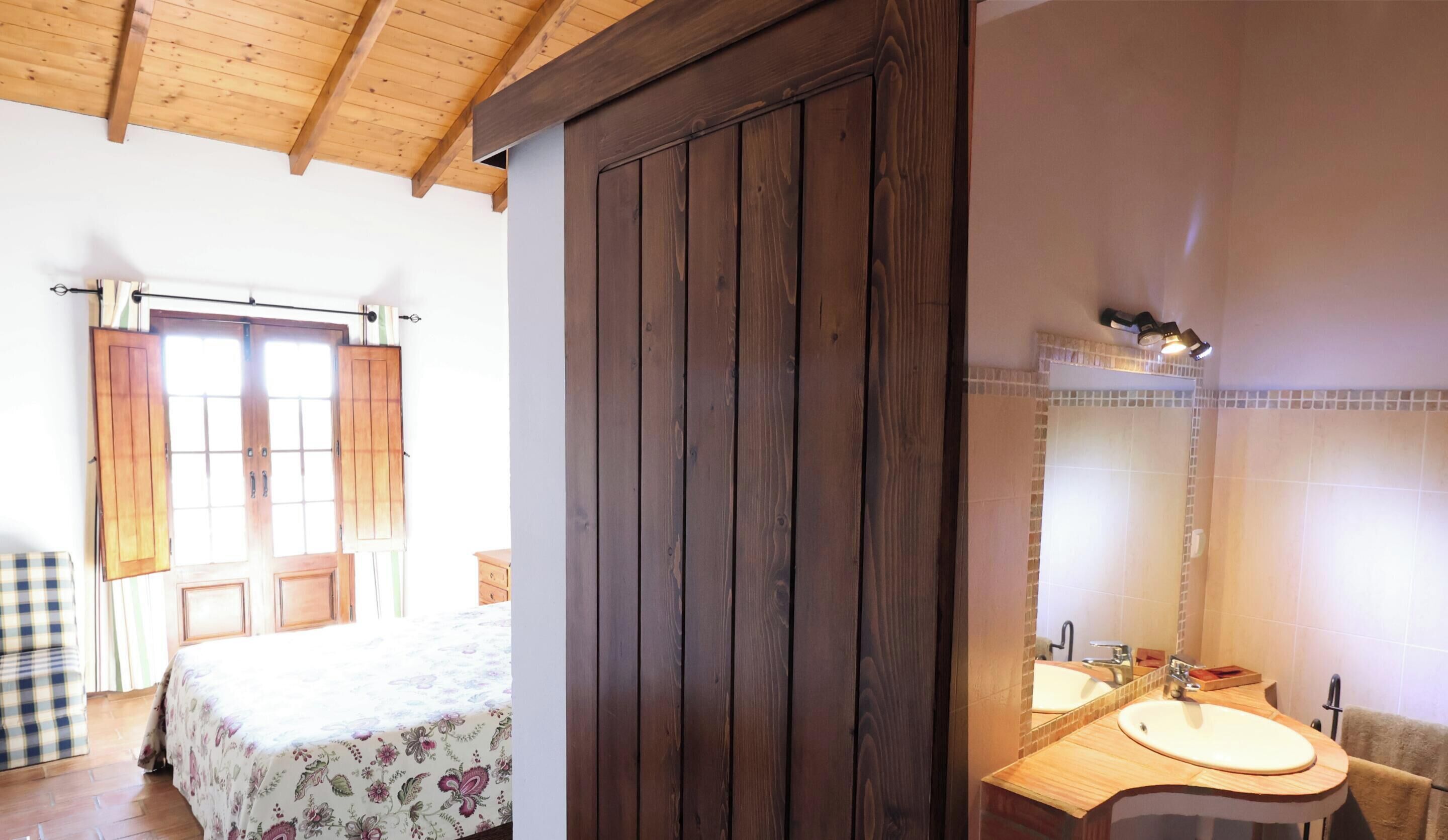 A room with a bed, a wooden wardrobe, a bathroom with a sink and mirror.
