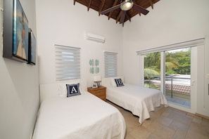 Ein Zimmer mit zwei Betten, einem Deckenventilator, einer Klimaanlage, einem Fenster mit Jalousien und Blick auf Grünfläche.