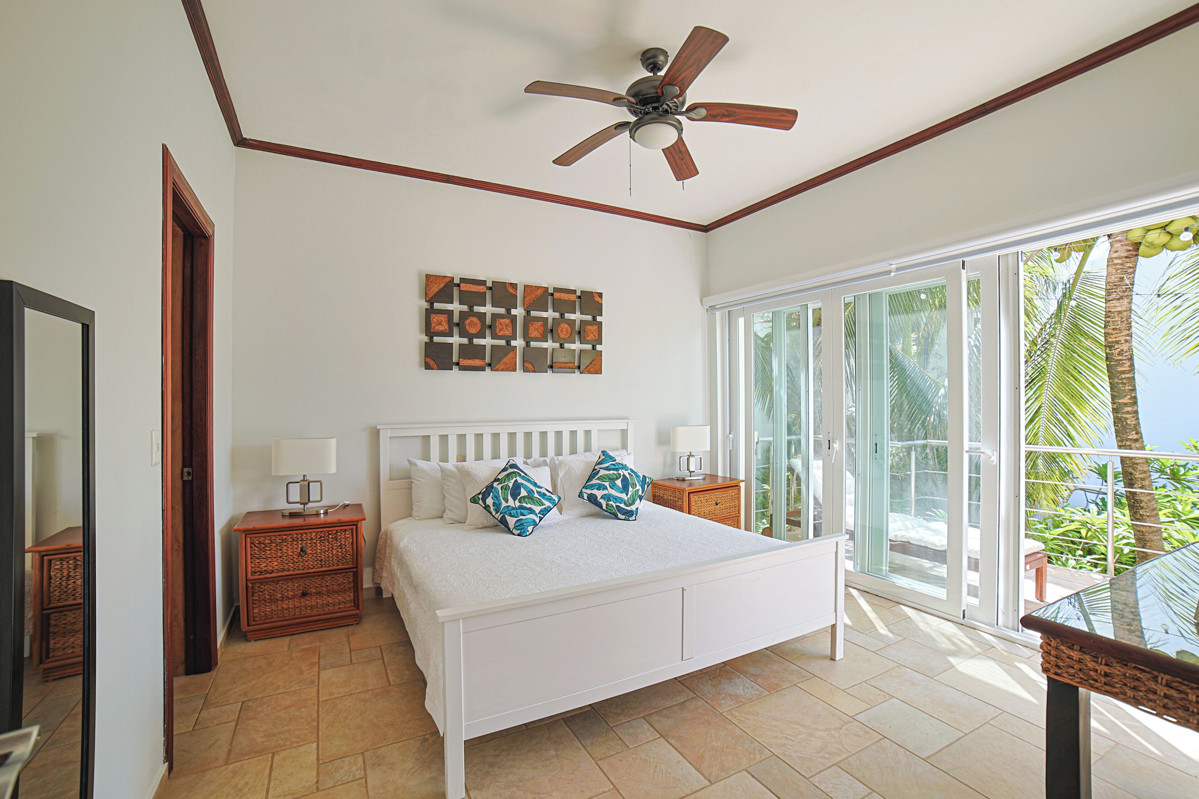 A bedroom with a bed, a ceiling fan, a window with a view of palm trees, and a wall with a geometric art piece.