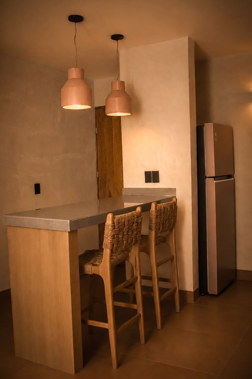 A compact kitchen with a central island, two bar stools, pendant lighting, and a built-in refrigerator.