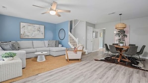 A modern living room with a sectional sofa, a small white table, a coffee table, and a dining area with a wooden table and chairs. - Chucktown Charm- 8 Mins to Credit One (Charleston)