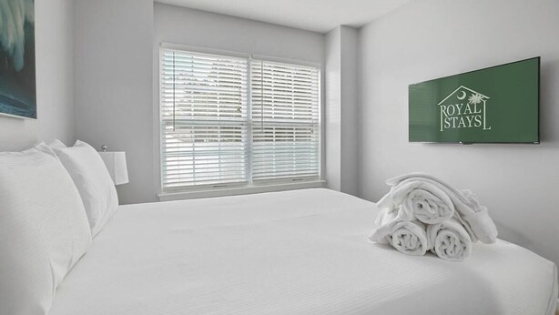 A hotel room with a neatly made bed, a window with blinds, a wall-mounted television, and a towel stack. - Chucktown Charm- 8 Mins to Credit One (Charleston)