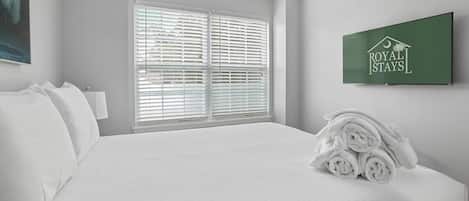A hotel room with a neatly made bed, a window with blinds, a wall-mounted television, and a towel stack.
