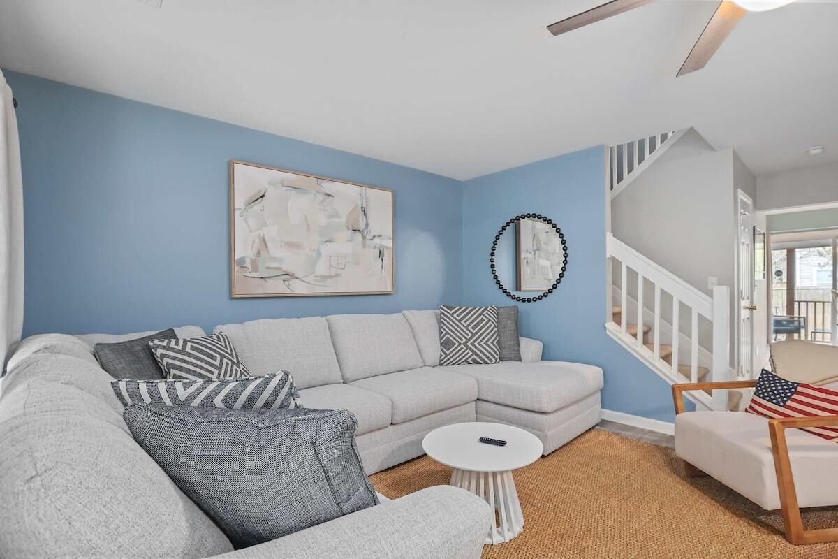 Ein Wohnzimmer mit einem hellgrauen Ecksofa, einem kleinen weißen Couchtisch und einem weißen Sessel mit Kissen mit amerikanischer Flagge. An der Wand hängt ein gerahmtes abstraktes Gemälde und darüber eine dekorative Spiegel.