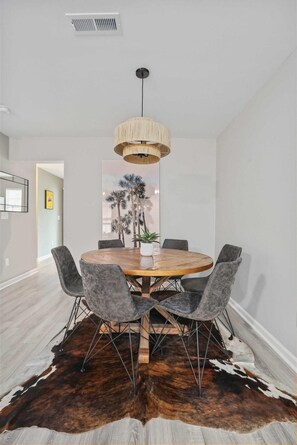 A dining area with a round wooden table, six grey upholstered chairs, a hanging light fixture, and a cowhide rug. - Chucktown Charm- 8 Mins to Credit One (Charleston)