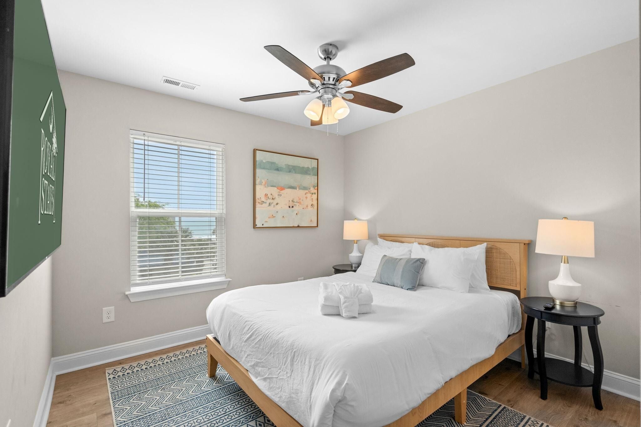 A bedroom with a bed, two bedside tables, a ceiling fan, a window with blinds, and a painting on the wall.