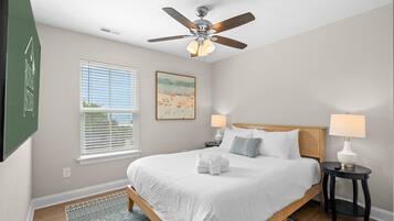 A bedroom with a bed, two bedside tables, a ceiling fan, a window with blinds, and a painting on the wall.