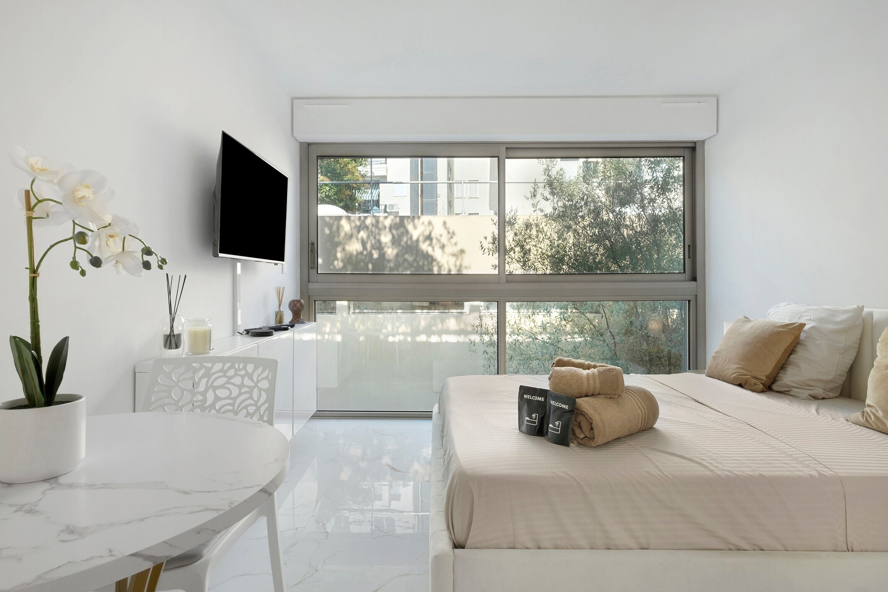 A modern hotel room with a large window, a bed with beige bedding, a small table with a potted plant, and a flat-screen TV mounted on the wall.