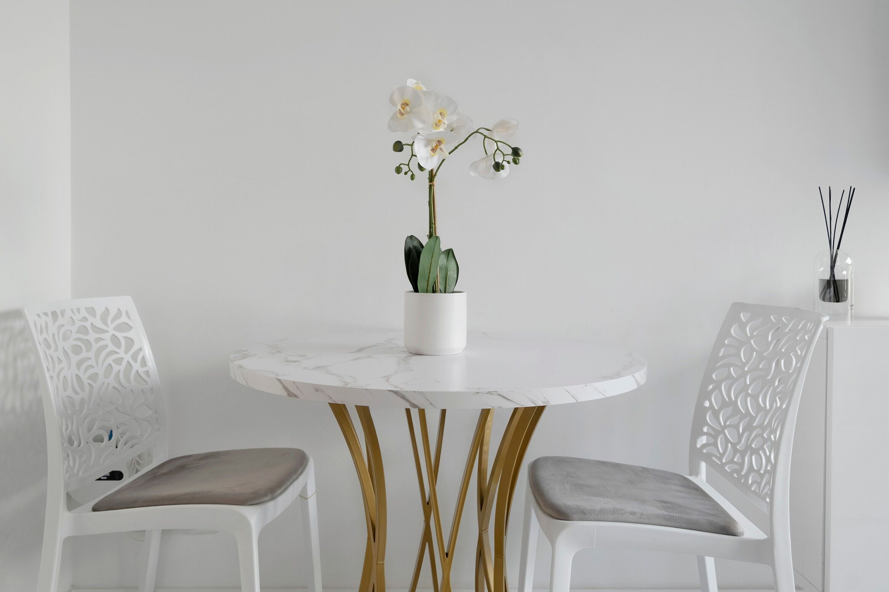 Una mesa redonda con tapa de mármol blanco, dos sillas con patas doradas y una orquídea blanca en maceta blanca.