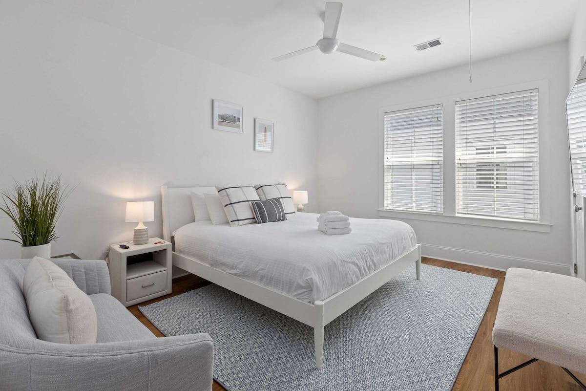 Une chambre moderne avec un lit blanc, des tables de chevet, un ventilateur de plafond et de grandes fenêtres équipées de stores.