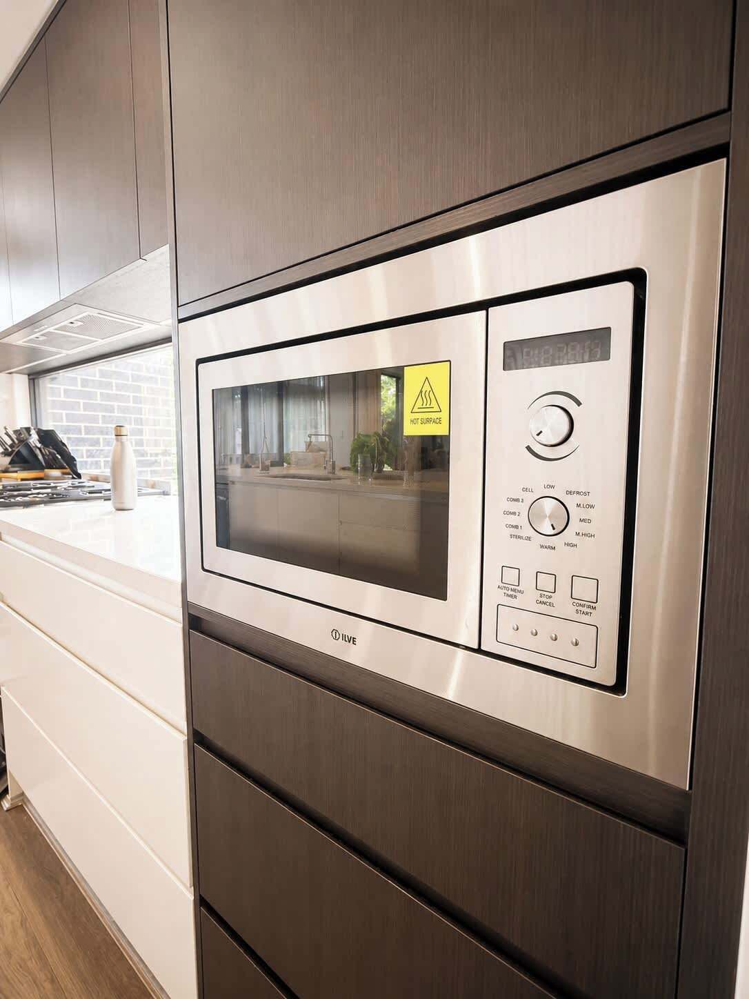A stainless steel microwave oven with a digital display and control panel, set in a modern kitchen with wooden cabinets and a window.