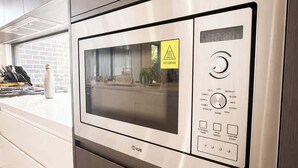 Forno de microondas em aço inoxidável com visor digital e painel de controle, inserido em uma cozinha moderna com armários de madeira e uma janela.