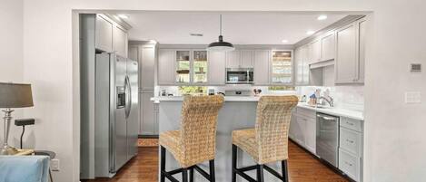 A modern kitchen with stainless steel appliances, a central island with bar stools, and a view into the adjacent living area.