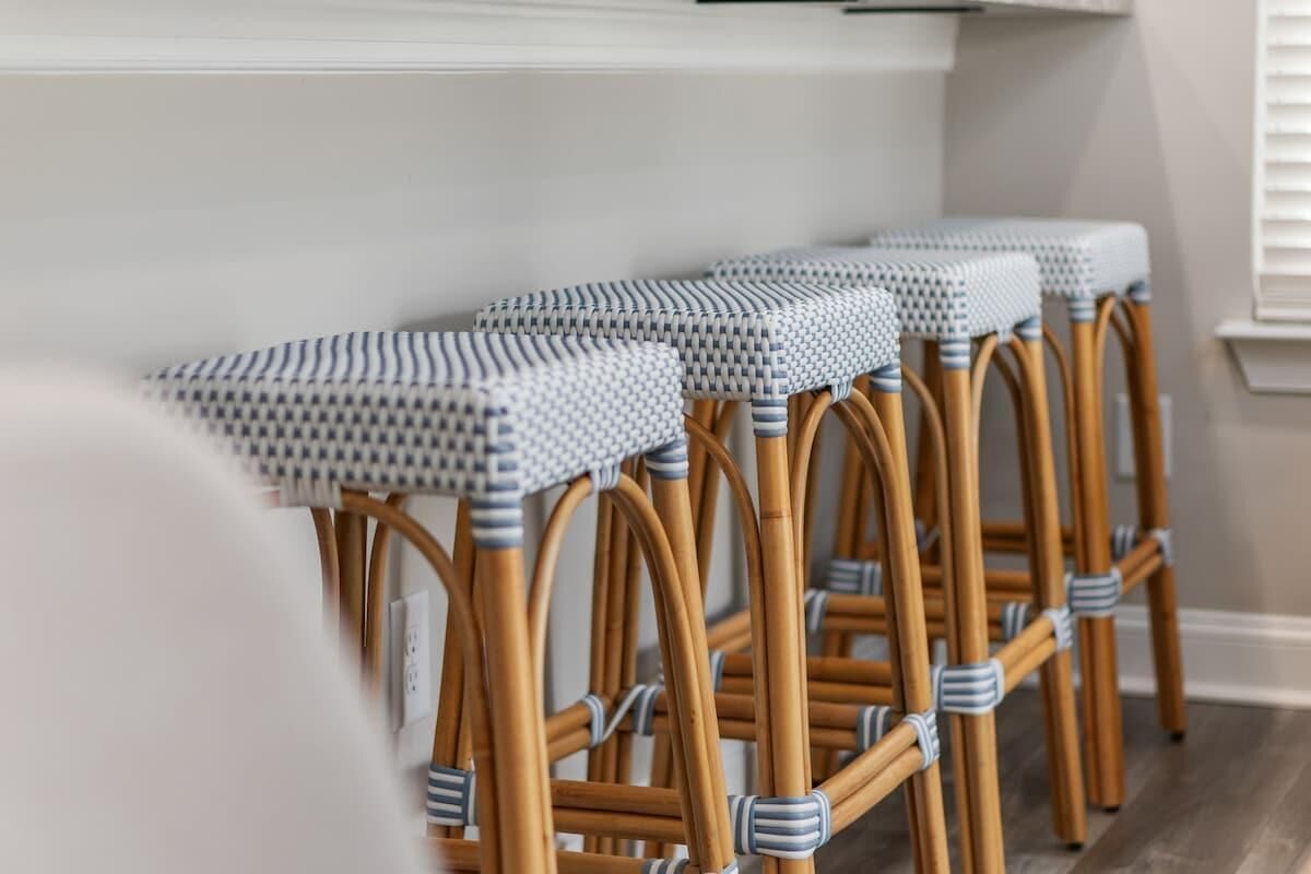 A row of wicker bar stools with woven tops and metal bands, positioned against a wall with a white baseboard and a window with blinds.