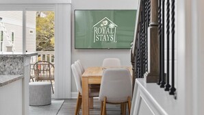 Ein Essbereich mit einem Holztisch und Stühlen, einem Fernseher, der 'ROYAL STAYS' zeigt, und Blick auf ein weißes Gebäude im Freien.