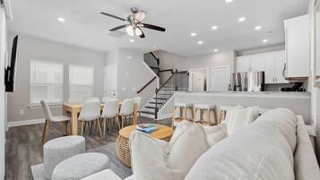 A modern living room with a sofa, chairs, a coffee table, and a dining area with a table and chairs. There is a kitchen area with cabinets and a refrigerator.