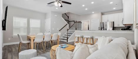 A modern living room with a sofa, chairs, a coffee table, and a dining area with a table and chairs. There is a kitchen area with cabinets and a refrigerator.