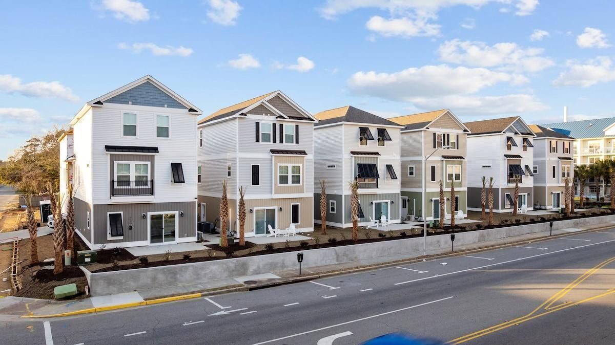 A row of modern, two-story townhouses with balconies, a paved driveway, and a clear sky.