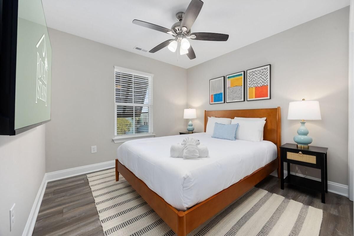 A hotel room with a bed, a ceiling fan, a window with blinds, a bedside table with a lamp, and artwork on the wall.