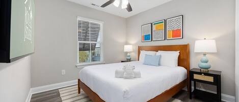 A hotel room with a bed, a ceiling fan, a window with blinds, a bedside table with a lamp, and artwork on the wall.