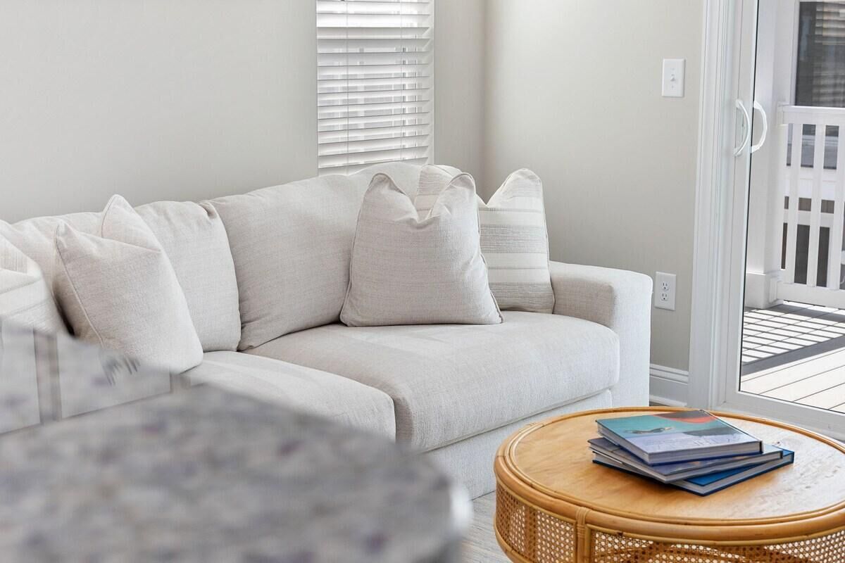 A living room with a light-colored sofa, a round wicker coffee table with books, and a view through a glass door to an outdoor balcony.