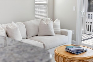 A living room with a light-colored sofa, a round wicker coffee table with books, and a view through a glass door to an outdoor balcony. - Sea Cloud-across Ocean-pet Friendly! (Myrtle Beach)