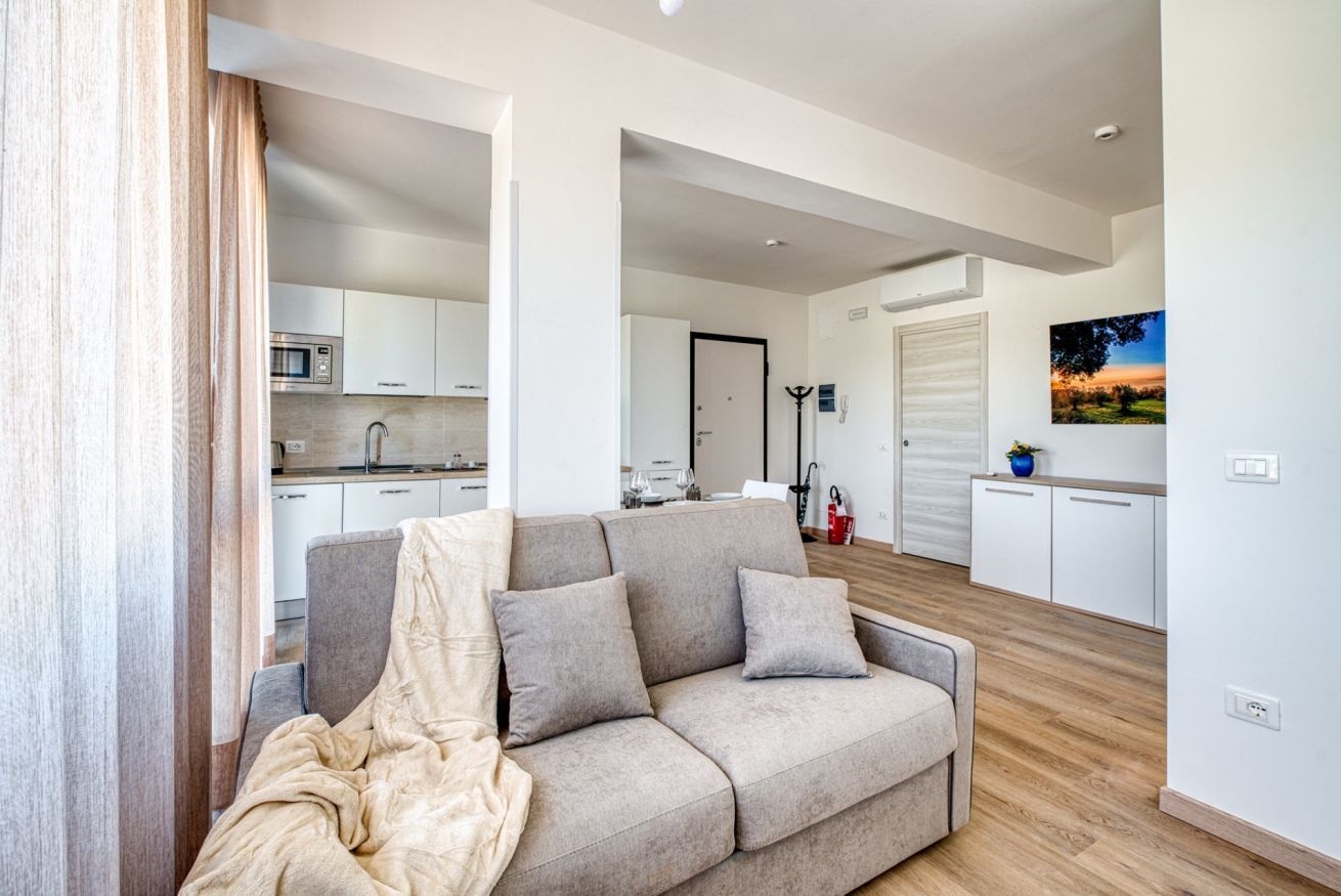 Een moderne woonkamer met een bank, een keuken op de achtergrond en een deur die naar een andere ruimte leidt.
