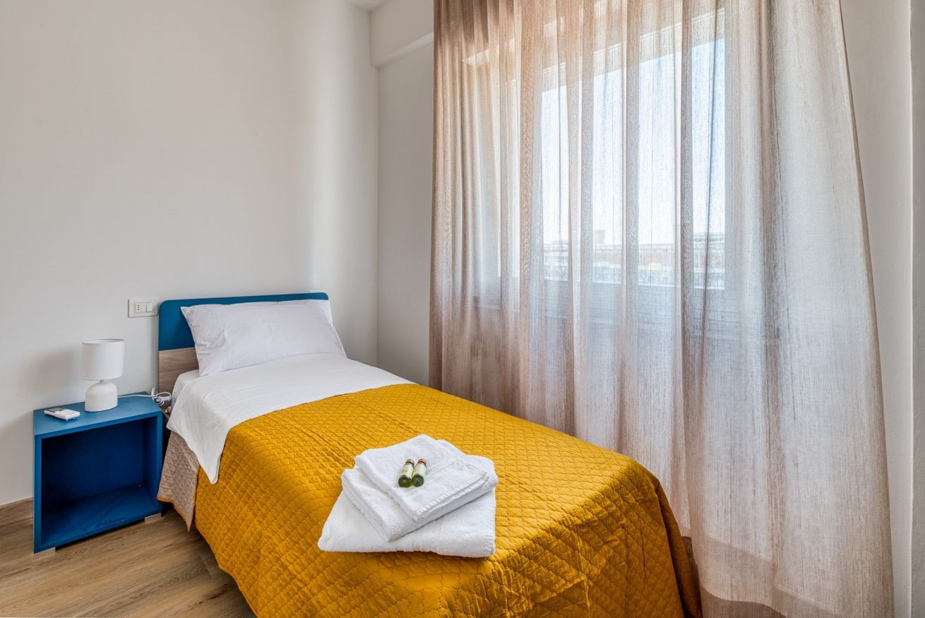 A hotel room with a bed, a nightstand, a lamp, and a window with sheer curtains.