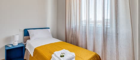 Habitación de hotel con una cama, una mesita de noche, una lámpara y una ventana con cortinas translúcidas.