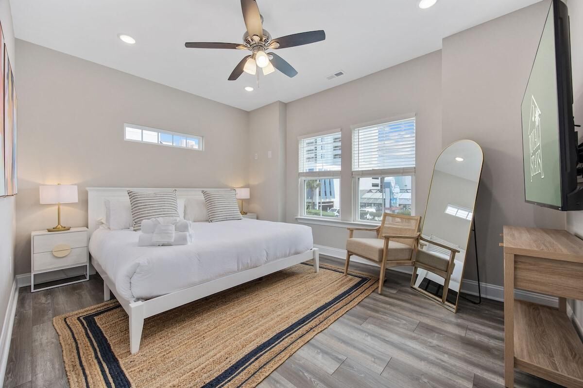 A bedroom with a bed, bedside tables, a ceiling fan, a mirror, and a window with blinds.