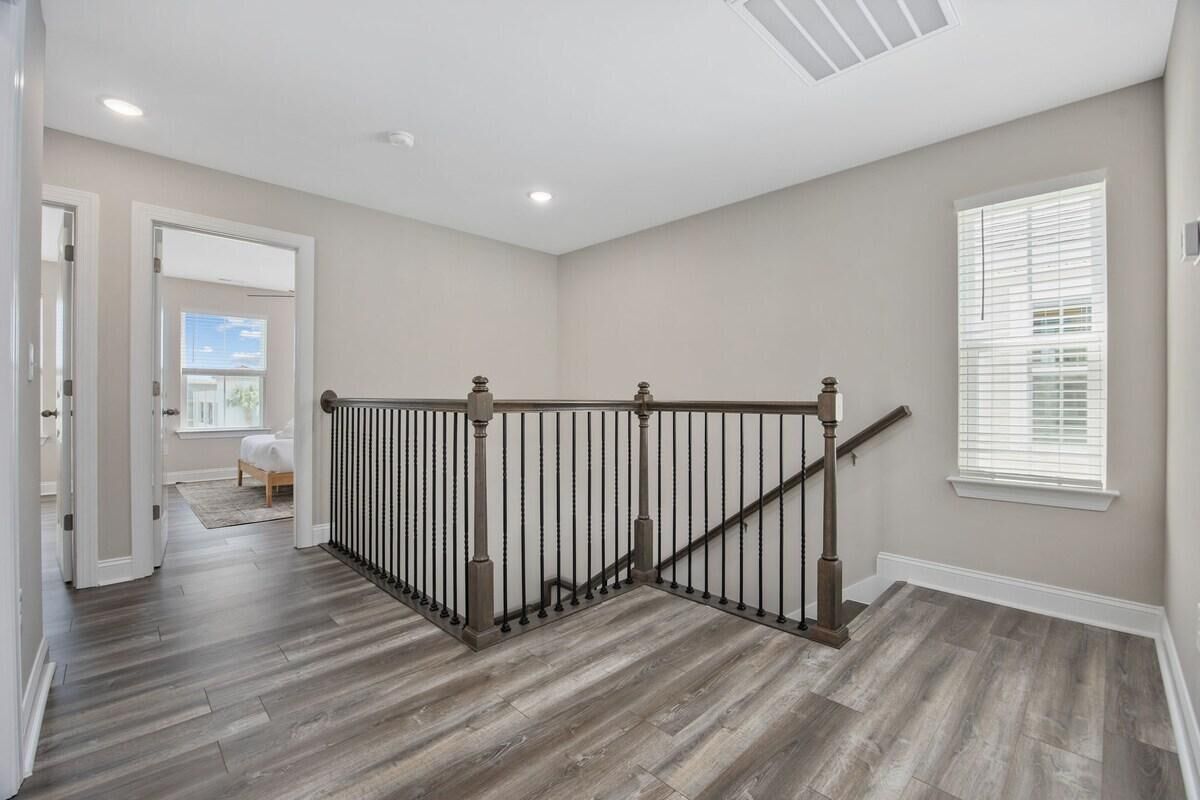 A spacious hallway with a wooden floor, a staircase with a metal railing, and a large window with blinds.
