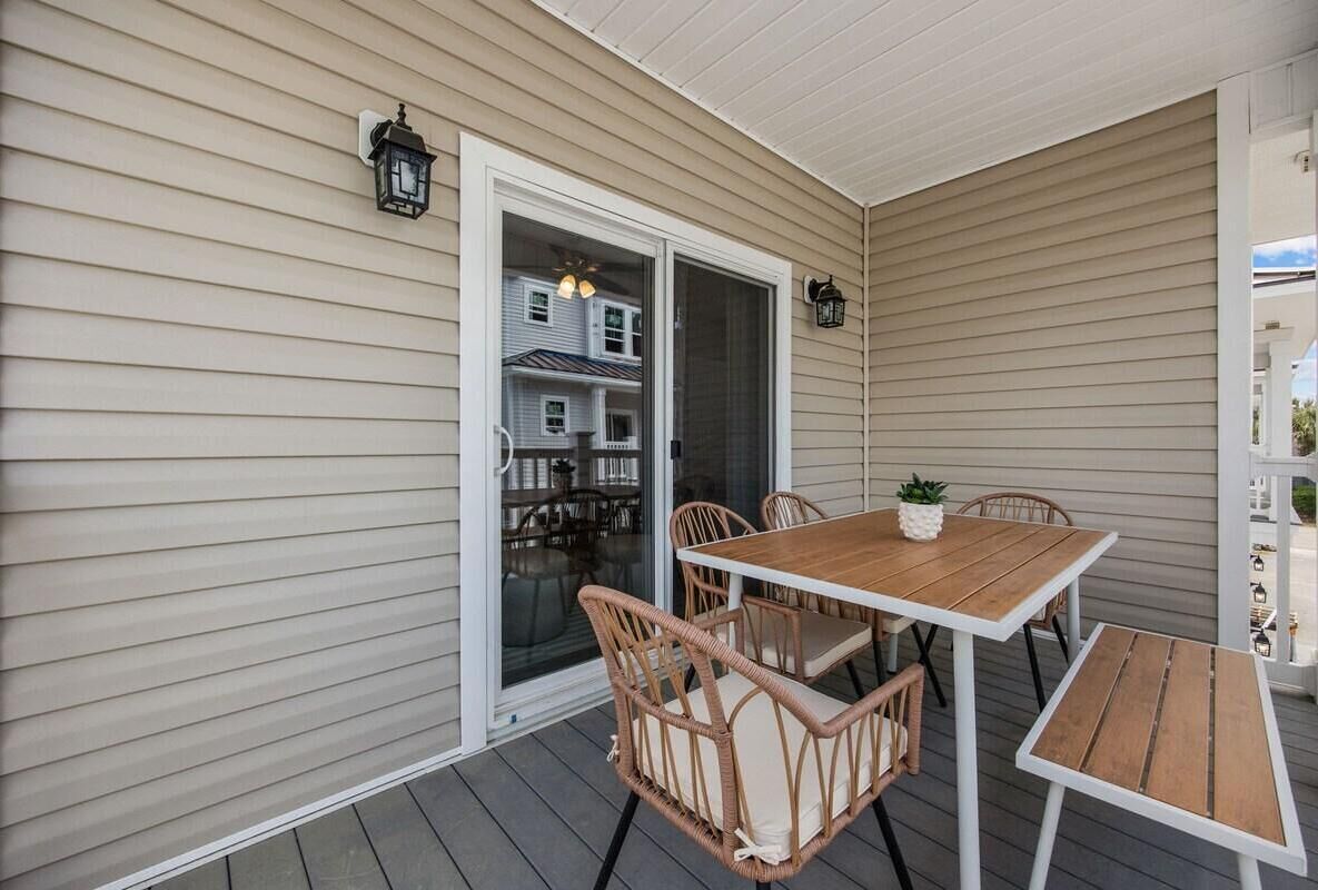 Une terrasse couverte avec une table en bois et des chaises en osier, une porte-fenêtre coulissante donnant sur un balcon, et deux lanternes fixées au mur.