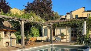 A swimming pool with a pergola and outdoor seating, surrounded by greenery and buildings. - Pool villa with panoramic view in Cortona (Sodo)