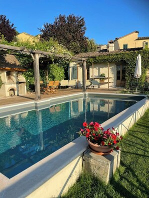 Ein Schwimmbecken mit einer Pergola und Sitzgelegenheiten im Freien, umgeben von Grünflächen und Gebäuden.