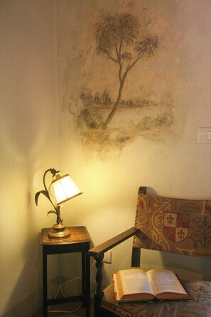Ein Zimmer mit einer Lampe auf einem kleinen Tisch, einem offenen Buch auf einem Stuhl und einem Wandbild mit einem Baum.