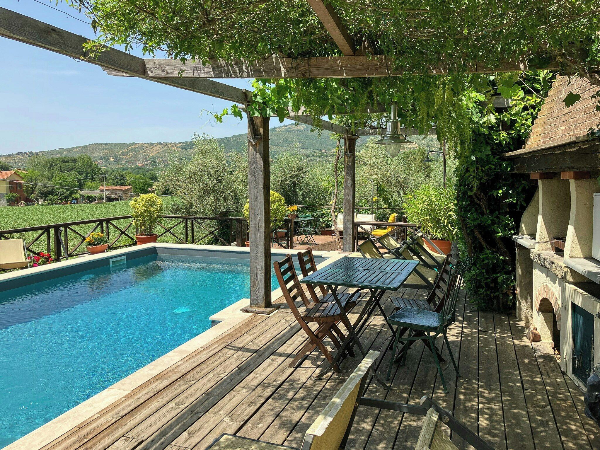 Eine Holzterrasse mit Pool, Gartenmöbeln und einer mit Weinreben bewachsenen Pergola.