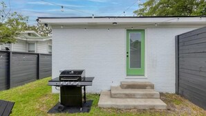 Une petite maison de plain-pied, à l ’extérieur blanc et à la porte verte. Devant la maison, on trouve un gril noir posé sur un tapis noir.