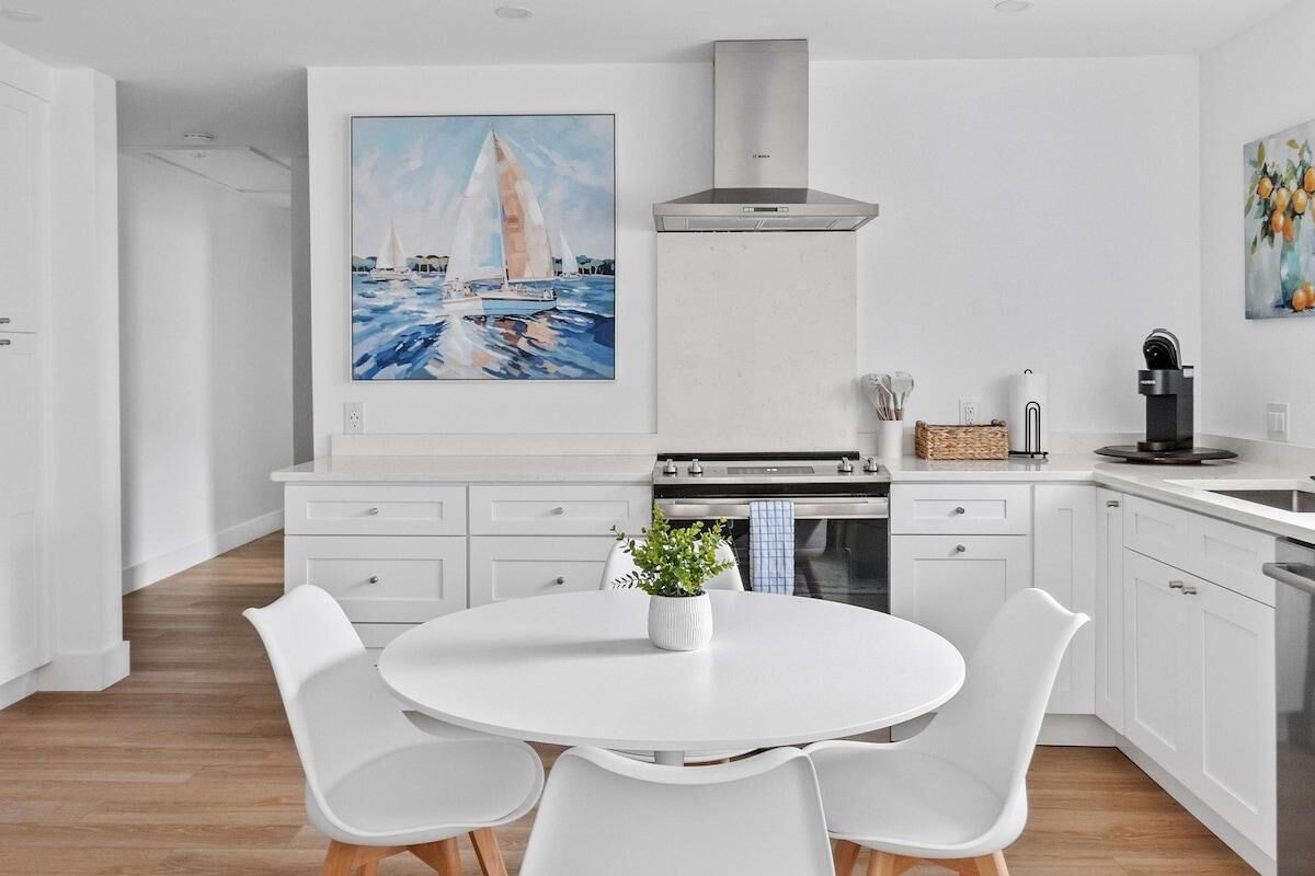 A modern kitchen with white cabinetry, a central island with chairs, a painting of sailboats, and a stainless steel range hood.