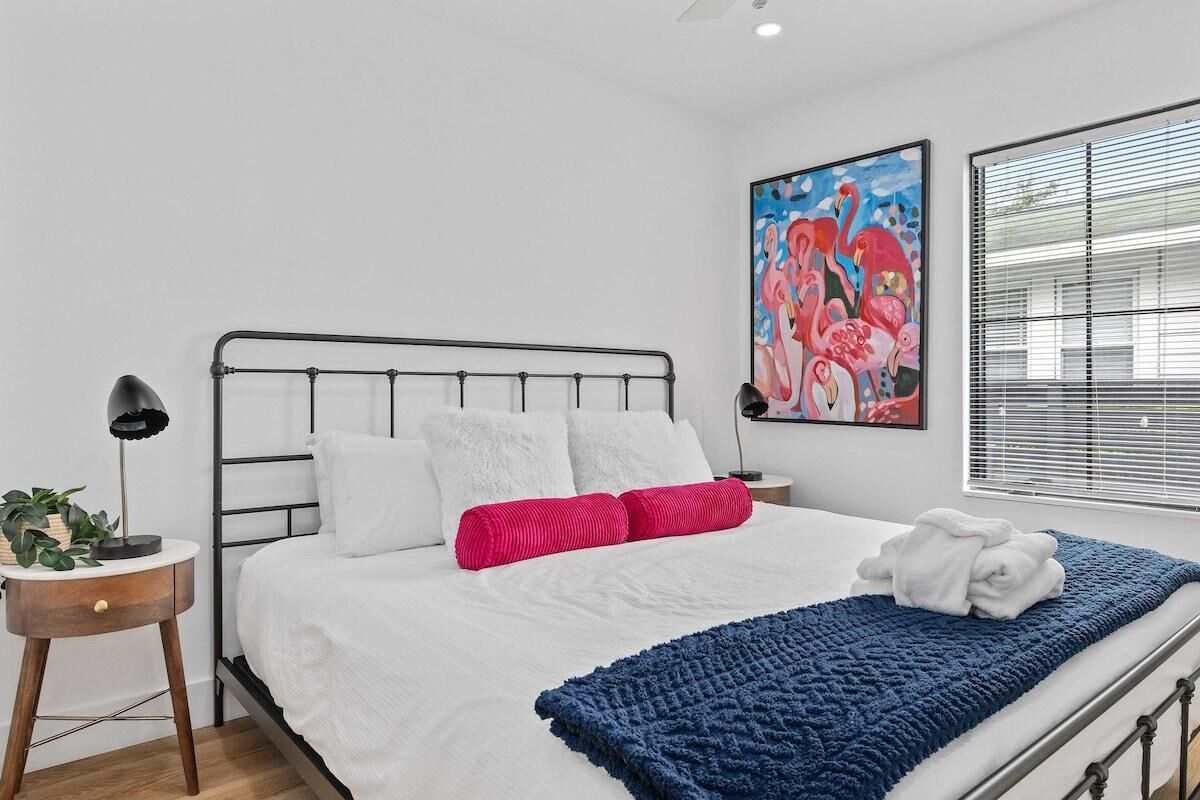 Un dormitorio moderno con estructura de cama metálica, ropa de cama blanca, una manta azul y una almohada rosa. Hay un cuadro en la pared y una ventana con persianas.