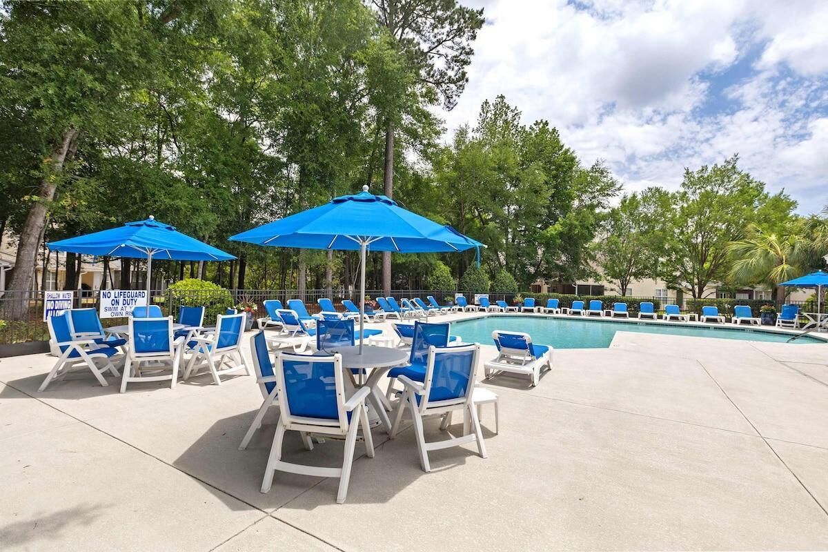 Un espace aménagé au bord de la piscine, avec des parasols bleus, des chaises longues blanches et un panneau indiquant qu’il n’y a pas de sauveteur de service.