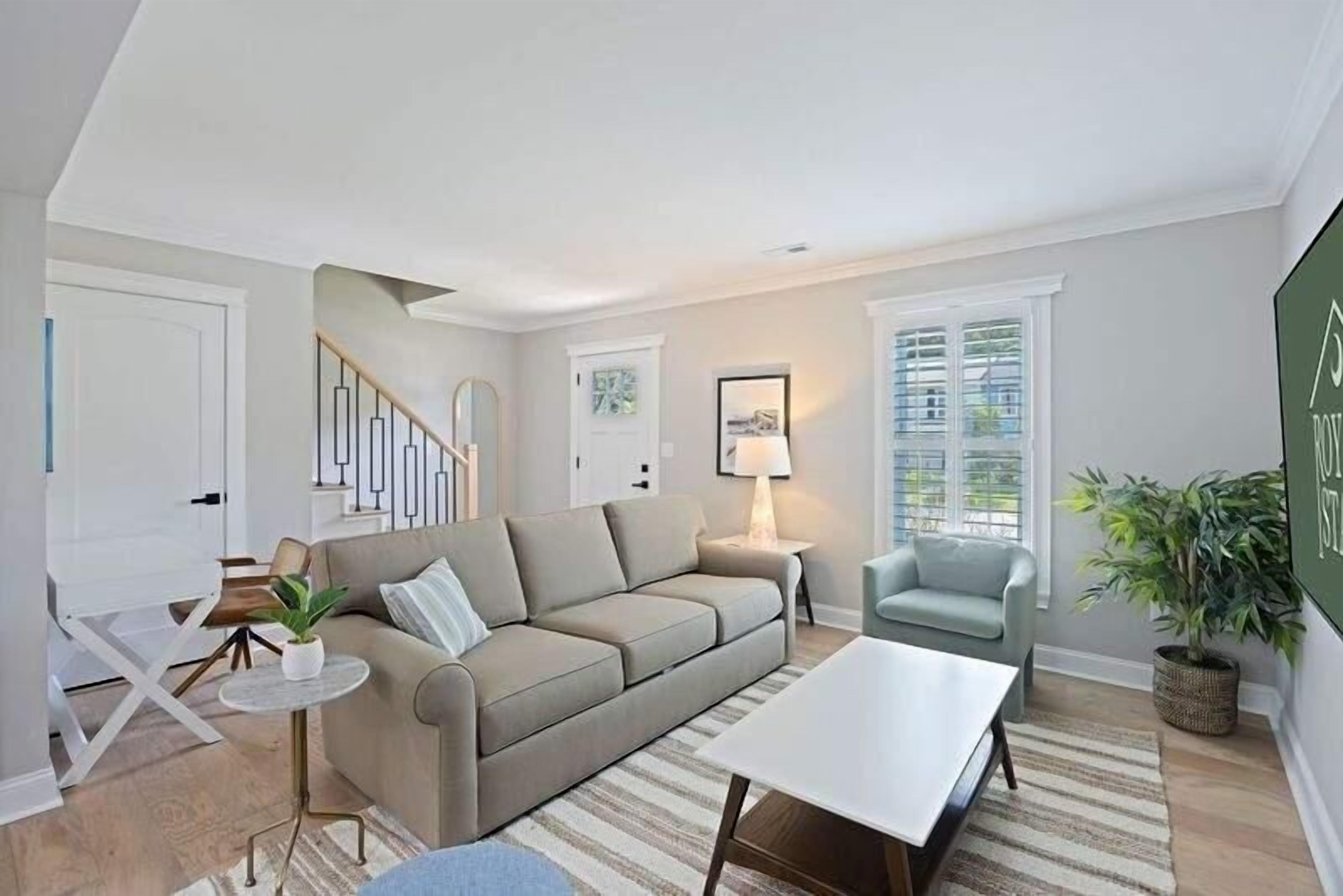A modern living room with a beige sofa, a white coffee table, and a blue armchair. There is a staircase with a railing, a lamp, and a framed picture on the wall.