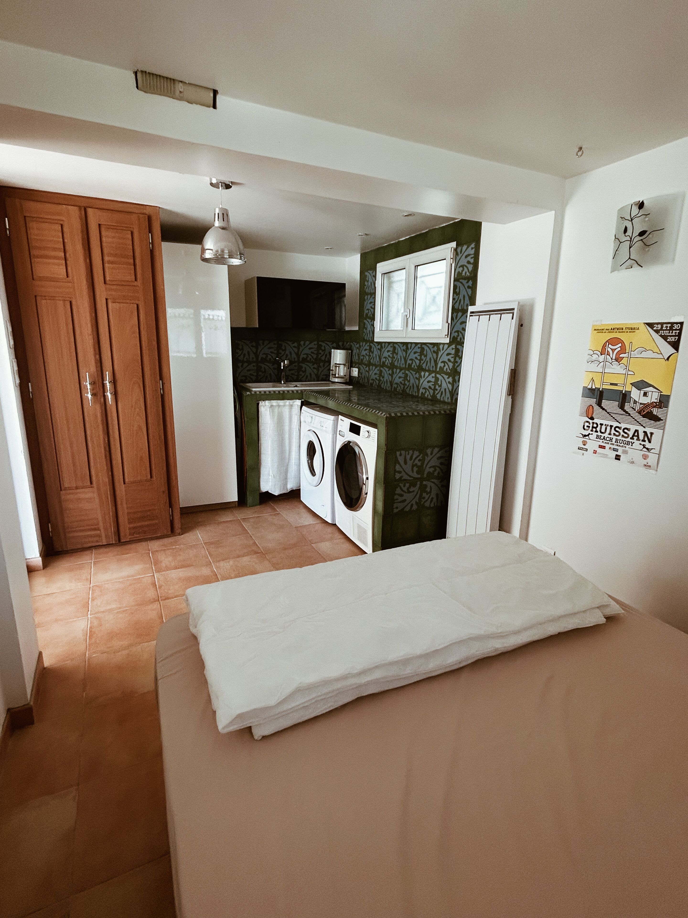 Une pièce de taille modeste comprenant un lit, un lave-linge, un évier et un petit coin cuisine.