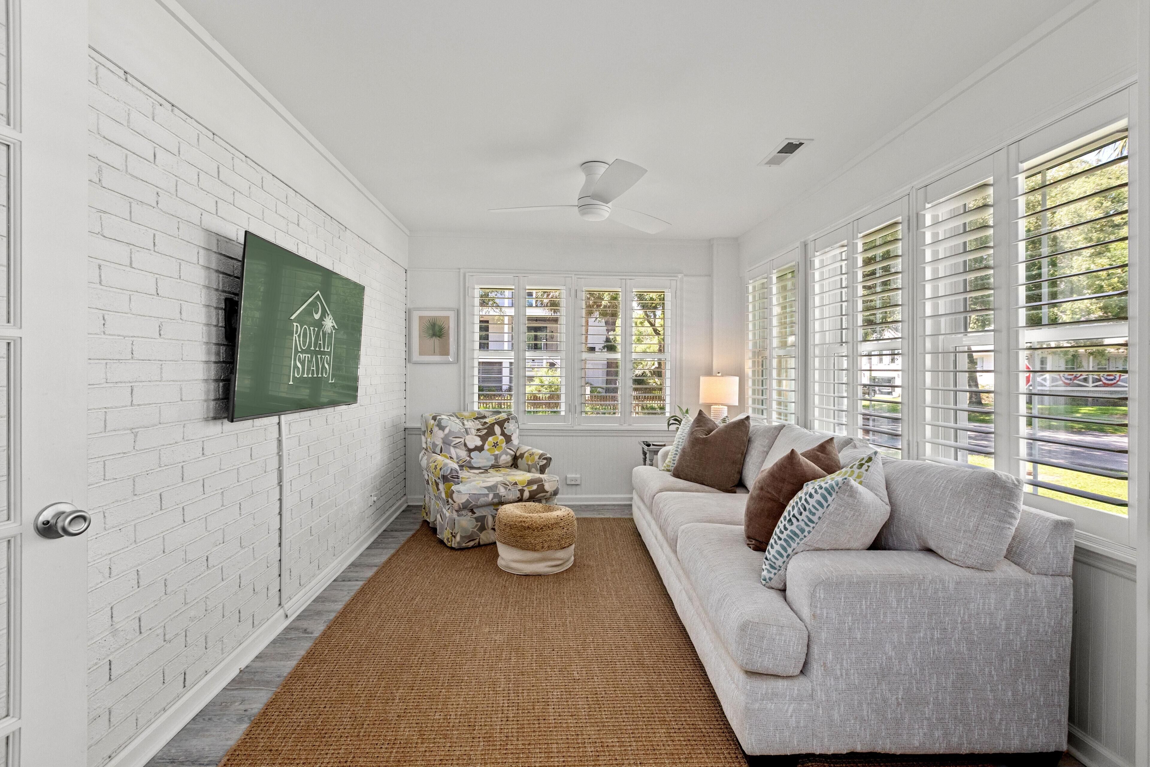Un salon avec un mur en briques blanches, un téléviseur à écran plat, un fauteuil à motifs floraux, un canapé gris et une fenêtre avec des stores.
