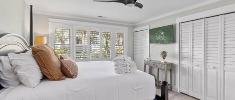 A bedroom with a bed, a ceiling fan, a window with blinds, a wall-mounted TV, and a wardrobe.