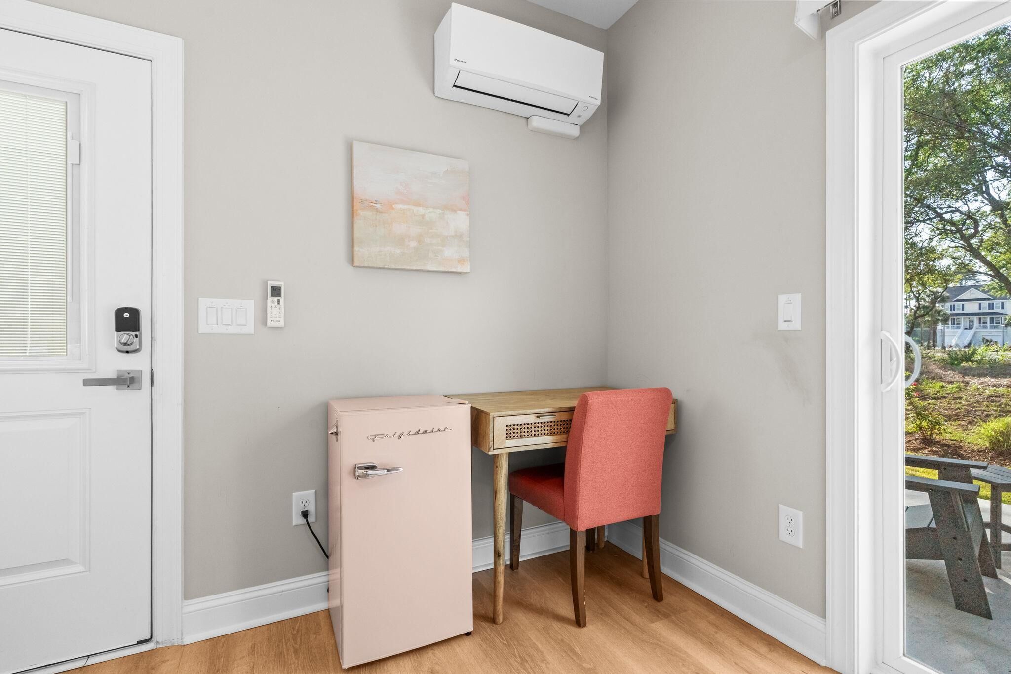 A room with a white door, a small pink refrigerator, a wooden desk with a red chair, a wall-mounted air conditioner, and a painting.