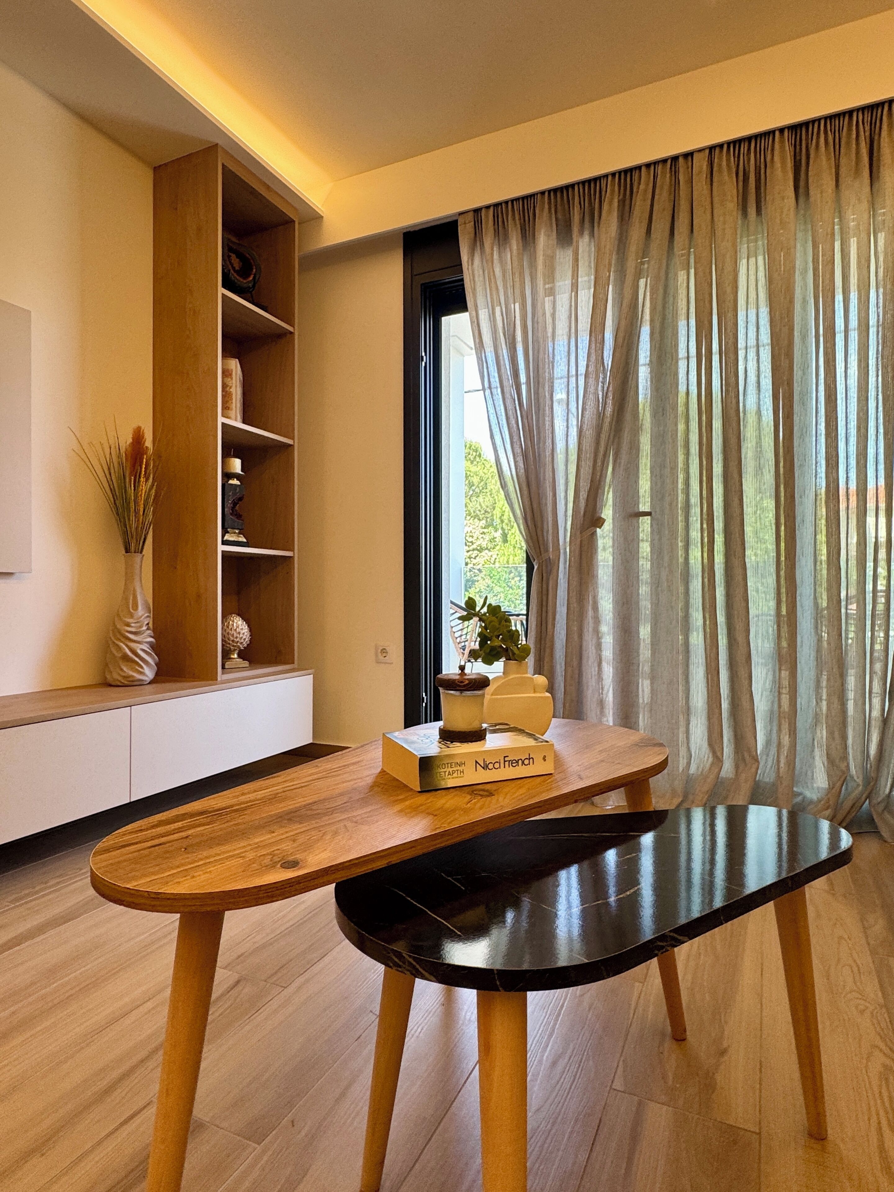 A modern living room with a wooden coffee table, a bookshelf, and a window with sheer curtains.